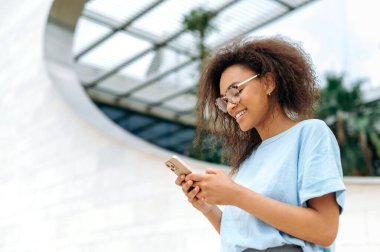 Online chatting. Positive african american curly haired young woman, stylishly dressed, using her smartphone while standing outdoors, messaging with friends in social networks, answers email, smiling
