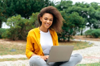 Positive trendy african american curly haired millennial girl, female student, freelancer, in stylish casual clothes, sitting with a laptop outdoors while online work or study, looks at camera, smiles