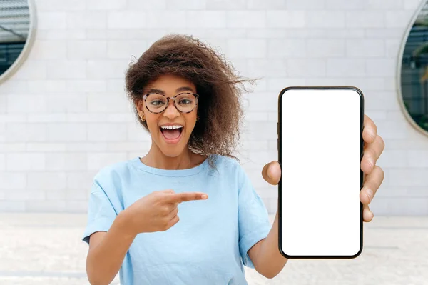 Empty space for advertising. Excited shocked amazed african american woman, stand outdoors, shows smartphone with blank white mock-up screen, points finger at it, looks surprised at camera