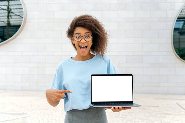 Excited amazed african american curly haired young woman, stands outdoors, holds an open laptop with blank white mock-up screen in hand, points finger at it, looks surprised at camera