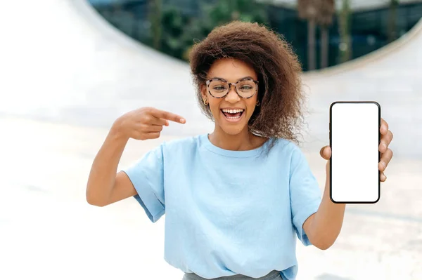 Mock-up concept. Excited mixed race young curly woman with glasses, stands outdoors, shows smartphone with blank white mock-up screen, points finger at it, looks amazed at camera, smiling happily
