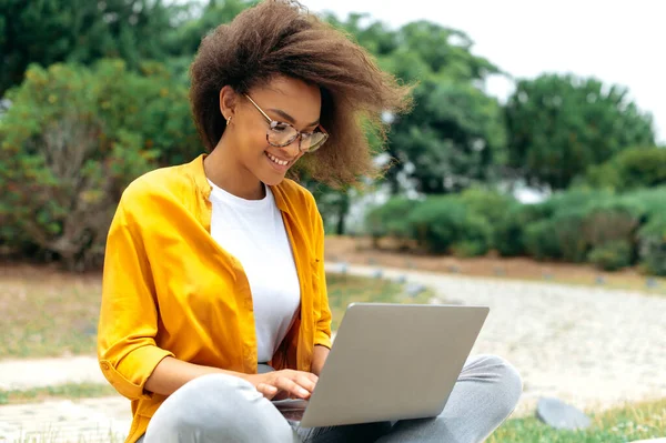 Busy successful african american curly haired young woman, student girl, freelancer, in stylish casual clothes, sit with laptop outdoors while online work or study, looking for information in internet
