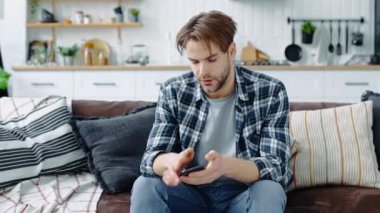 Kafası karışmış, kafası karışmış, beyaz bir genç adam, oturma odasında bir kanepede oturuyor, akıllı telefon ekranına bakıyor, başarısız proje, kötü haber ya da mesaj alıyor, umutsuzluk duyguları, kişisel problemler.