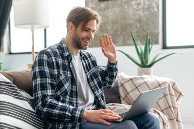 Yakışıklı beyaz adam internette kanepe sohbetleri yapıyor. Dizüstü bilgisayar kullanan çekici serbest çalışan, video görüşmesi yapan, iş arkadaşlarıyla video konferansı yapan, el sallayan ve gülümseyen biri.
