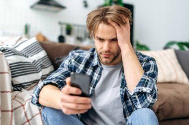Hayal kırıklığına uğramış Kafkasyalı genç bir adam, oturma odasındaki kanepenin yanında oturuyor, üzgün bir şekilde akıllı telefon ekranına bakıyor, yalnızlığı hissediyor, kötü haber ya da mesaj alıyor, umutsuzluk duyguları yaşıyor.