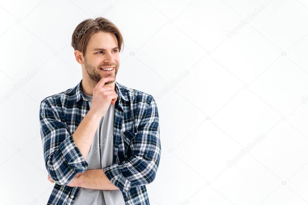 Photo of an attractive confident caucasian stylish young man, in casual clothes, standing on a white isolated background, looking to the side, thinking, smiling friendly. Space for presentation
