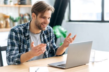 Video iletişimi. Kafkasyalı arkadaş canlısı genç bir adam, şirket çalışanı, serbest çalışan, mutfakta oturan, bilgisayarını uzaktan kullanan, müşteri ya da iş arkadaşıyla video konferansında konuşan, gülümseyen