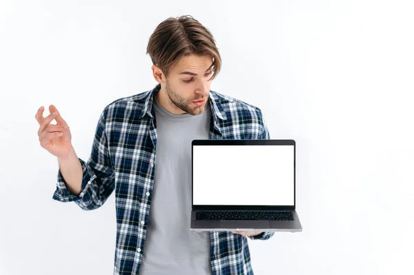 Mock-up concept. Confused puzzled caucasian guy in casual wear, freelancer or student, holding an open laptop with empty mock-up space, peeks at it in surprised, stand on white isolated background