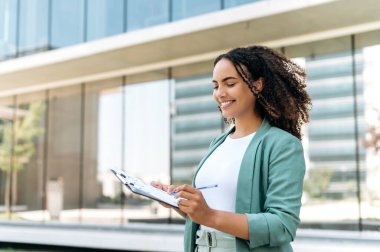 Güzel, kendine güvenen başarılı Latin ya da Brezilyalı kıvırcık saçlı, iş kadını, finans uzmanı, şık giyinen, açık havada mali tablolarla duran, grafikleri analiz eden, gülümseyen