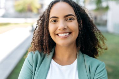 Güzel, kendine güvenen, başarılı İspanyol ya da kıvırcık saçlı, iş kadını, şık şık elbiseli, dışarıda duran, kameraya bakan, gülümseyen güzel ve kendine güvenen genç bir kadının yakın çekimi.