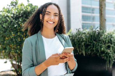 Cihazı kullanarak. Pozitif İspanyol ya da Brezilyalı kıvırcık saçlı, şık giyimli, cep telefonunu kullanarak dışarıda duruyor, sosyal medyada arkadaşlarıyla mesajlaşıyor, kameraya bakıyor, gülümsüyor.