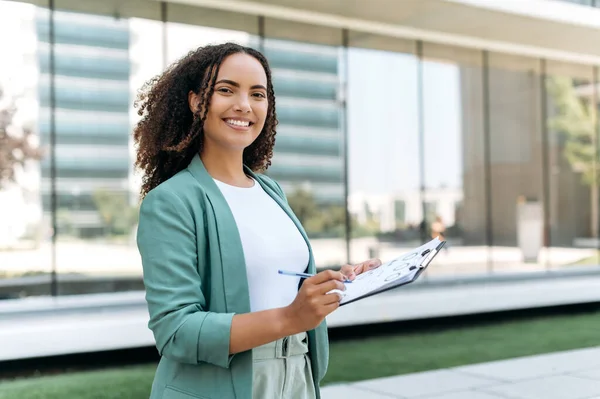 Portrait of lovely positive confident successful mixed race young woman with curly hair, business lady, trading broker, in elegant wear, standing outdoors with financial charts, looks at camera, smile