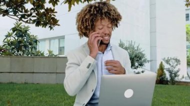 Positive busy successful hispanic or brazilian business man or top manager, works outdoors in laptop, talking on smartphone with client or colleague, typing on keyboard. Phone conversation during work