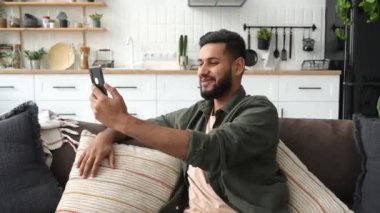 Positive mixed race young stylish man sitting on sofa in living room, using his smart phone, talking on video call. Happy indian or arabian video blogger, makes a selfie phone, records a stream, smile