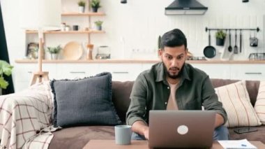 Cheerful amazed arabian or indian guy, freelancer, sits on a sofa in living room, looks at laptop screen, gesticulates with hands, celebrates success, victory, big profit, received good news, smiles