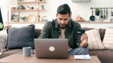Online payment. Indian or Arabian positive young male in casual wear, sits in cozy living room, holding bank card in hand, using laptop, shopping online, ordering home delivery, paying bills, smiling