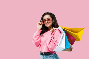 Alışveriş konsepti. Kafkasyalı şık, şık, esmer bir kadın, günlük kıyafetler ve pembe güneş gözlüğü takıyor, alışveriş torbaları tutuyor, pembe arka planda duruyor, kameraya bakıyor, gülümsüyor.