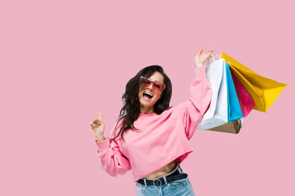 Shopping and sales concept. Cheerful trendy pretty caucasian young woman, in casual clothes and pink sunglasses, holding shopping bags, posing on isolated pink background, looks at camera, smiling