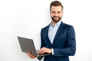 Arkadaş canlısı beyaz sakallı iş adamı, takım elbiseli, programcı, bilişim uzmanı, dizüstü bilgisayarı elinde tutuyor, izole edilmiş beyaz arka planda duruyor, kameraya bakıyor, gülümsüyor.