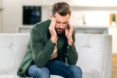 Stresli, mutsuz, beyaz bir adam, oturma odasında bir kanepede oturuyor, baş ağrısı çekiyor, migren, şakaklarına masaj yapıyor, gözlerini kapatıyor, sağlıksız hissediyor, aşırı çalışıyor, dinlenmeye ihtiyacı var.