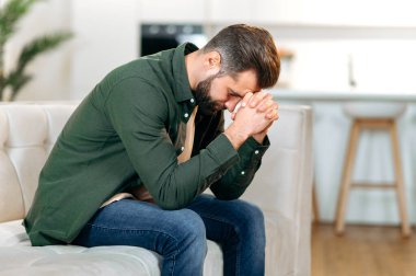 Kafkasyalı üzgün bir adam, oturma odasındaki kanepede oturuyor ve gözleri kapalı, kişisel problemler yaşıyor, stres, yalnızlık, psikolojik desteğe ve dinlenmeye ihtiyacı var.