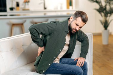 Kafkasyalı mutsuz bir adam, gündelik kıyafetler giymiş, oturma odasında kanepede oturuyor, sırtında ve sırtında şiddetli ağrılar var, belinde kronik eklem problemleri var. Sırtında, omurgasında, siyatik problemleri var.