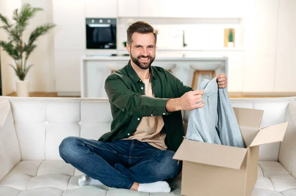 英俊积极的高加索男人坐在沙发上打开大箱子 打开家里的网店订单 对快递服务和收到的货物感到满意 开心地笑着 — 图库照片