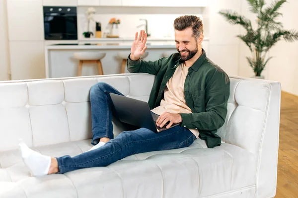 Positive handsome caucasian man, in stylish casual wear, having online video conference with friends or colleagues by laptop at home while sitting on comfortable sofa, waves hand, smiling friendly