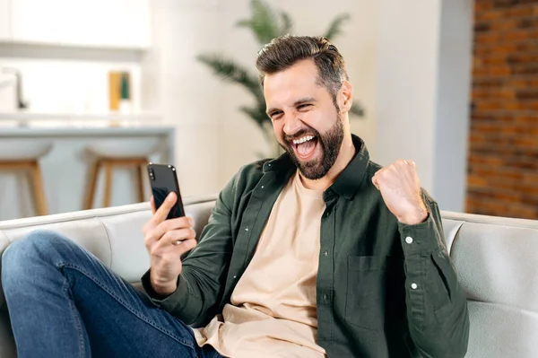 Joyful lucky attractive caucasian man in casual wear, sitting on a couch in cozy living room, using his smart phone, reading good message or news, gesturing with fist, smiling. Digital technology