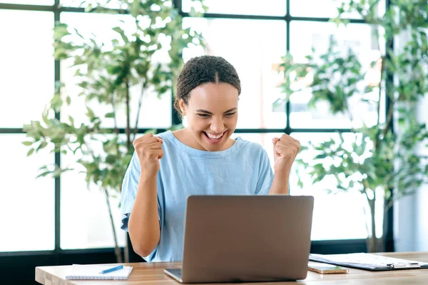 Win, success. Cheerful happy brazilian or hispanic young business woman, sits at a desk with a laptop in modern office, rejoicing in success, big profit, celebrate deal, gesturing with fists, smiling
