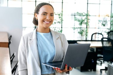 Güzel, zarif, başarılı, melez bir iş kadını, programcı, şık bir iş elbisesi içinde, ofiste duruyor, elinde açık bir dizüstü bilgisayarla, kameraya bakıyor, dostça gülümsüyor.