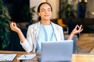 Pozitif sakinlik rahatlatıcı genç melez kadın, ofis çalışanı, şirket müdürü, ofiste oturan, işten dinlenen, kapalı gözlerle meditasyon yapan, gülümseyen.