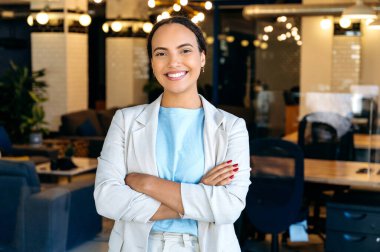 Kendine güvenen, başarılı, modern, melez bir iş kadınının portresi, modern yaratıcı ofisinde duran, resmi giyinmiş, kollarını kavuşturmuş, kameraya bakan, gülümseyen bir şirket yöneticisi.