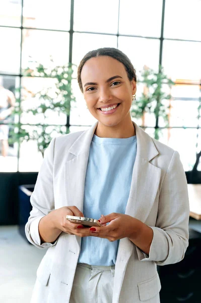 Online messaging. Positive mixed race business woman, standing in modern creative office, uses her smart phone, texting in social media, answers email, browsing internet, looks at camera, smiling