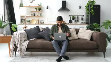 Tatmin olmuş Hintli ya da Arap genç adam, oturma odasında kanepede oturuyor, işini bitiriyor ya da laptopta çalışıyor, onu kapatıyor, ellerini başının arkasına koyup kanepeye uzanıyor, başka yere bakıyor, dinleniyor, gülümsüyor.