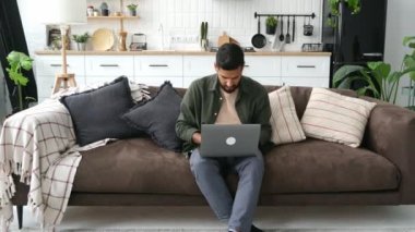 Hüsrana uğramış Arap ya da Hintli genç adam, oturma odasında kanepede oturuyor, laptopta çalışıyor ya da çalışıyor, stresli ve fazla çalışıyor, bilgisayarını kapatıyor, işten bıkmış, dinlenmeye ihtiyacı var.