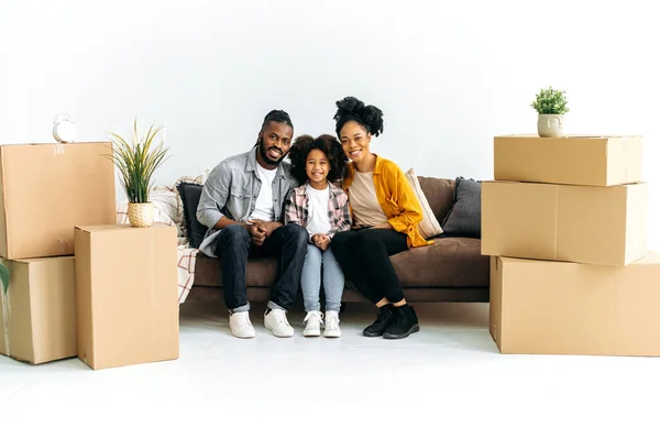 Family portrait of happy african american family, mom, dad and their little daughter spend time at home together, hugging on sofa between cardboard boxes in their new home, looking at camera, smiling