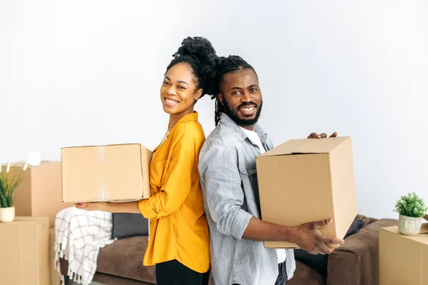 Happy modern married african american couple, holding boxes with things for the house, standing in their new housing, looking at the camera, smiling, rejoicing in buying their own apartment or house