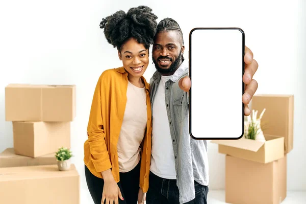 幸福的非洲裔美国夫妇 新婚夫妇 站在客厅里 背对着装有新家物品的纸盒 用空白的白色模拟屏幕展示智能手机 — 图库照片