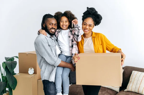 New housing, relocation. Happy african american family, dad mom and their cute daughter, moved to their new apartment, holding boxes with things, girl in dads arms shows keys, they smile into camera