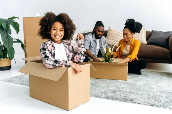 一个非裔美国人家庭搬进了他们的新家 快乐可爱的学龄前女孩 坐在客厅的盒子里 拿出钥匙 看着相机 微笑着 家长们正在整理一个盒子的背景 — 图库照片