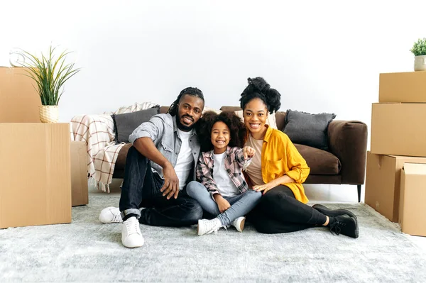 积极快乐的非裔美国人家庭搬进了他们的新家 妈妈爸爸和他们的女儿 坐在客厅纸板箱中间的地板上 女孩拿出钥匙 她们看着相机 微笑着 — 图库照片