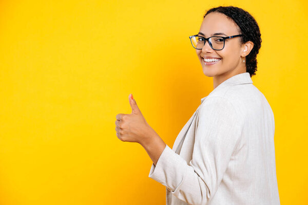 Счастливая успешная красивая бразилька или латиноамериканка деловая женщина, в повседневной куртке, стоит спиной к камере на изолированном оранжевом фоне, показывая большой палец вверх жестом, улыбается в камеру