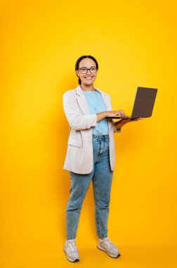 Pozitif özgüvenli İspanyol ya da Brezilyalı genç bayan, gözlüklü, elleri açık, dizüstü bilgisayarı olan, izole edilmiş turuncu arka planda duran, kameraya bakan, gülümseyen