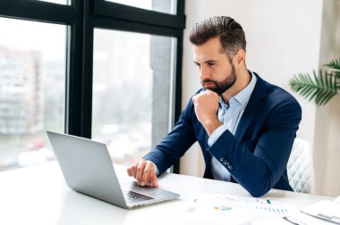 Odaklanmış başarılı, kendine güvenen, resmi takım elbiseli beyaz iş adamı, iş yerinde oturuyor, dizüstü bilgisayar kullanıyor, iş stratejisi yazıyor, bilgiyi analiz ediyor, internette geziniyor.