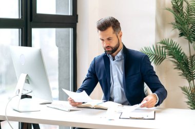 Seçkin, başarılı, beyaz iş adamı, şirket patronu, finans müdürü takım elbise giymiş, iş yerinde belgelerle çalışıyor, finansal grafikleri analiz ediyor, planlama stratejisi geliştiriyor.