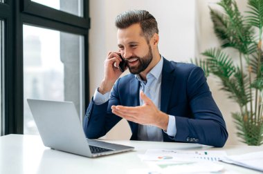 Takım elbiseli başarılı bir beyaz adam, şirket çalışanı, üst düzey yönetici, patron, ofiste oturuyor, iş arkadaşı veya müşterisiyle konuşuyor, iş sorunlarını çözüyor, gülümsüyor.