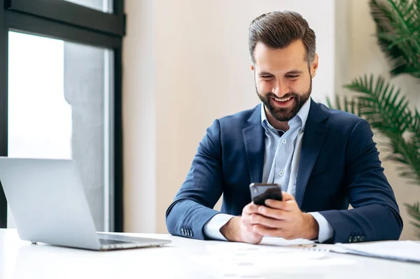 Online messaging. Positive busy caucasian business man in a suit, company employee, top manager, sitting in the office, using his smart phone, texting online with colleague or client, smiling