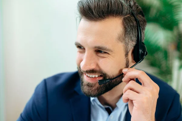 Close-up photo of a positive caucasian man with headset, call center worker, expert, company representative, support service operator, holds online consultation with client, looks to the side, smiling