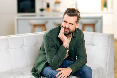 Diş ağrısı. Mutsuz, üzgün, üzgün, beyaz bir adam evdeki kanepede oturuyor, elini yanağına yaklaştırıyor, yüzünü buruşturuyor, dişleri ağrıyor, bir dişçiye görünmesi gerekiyor.